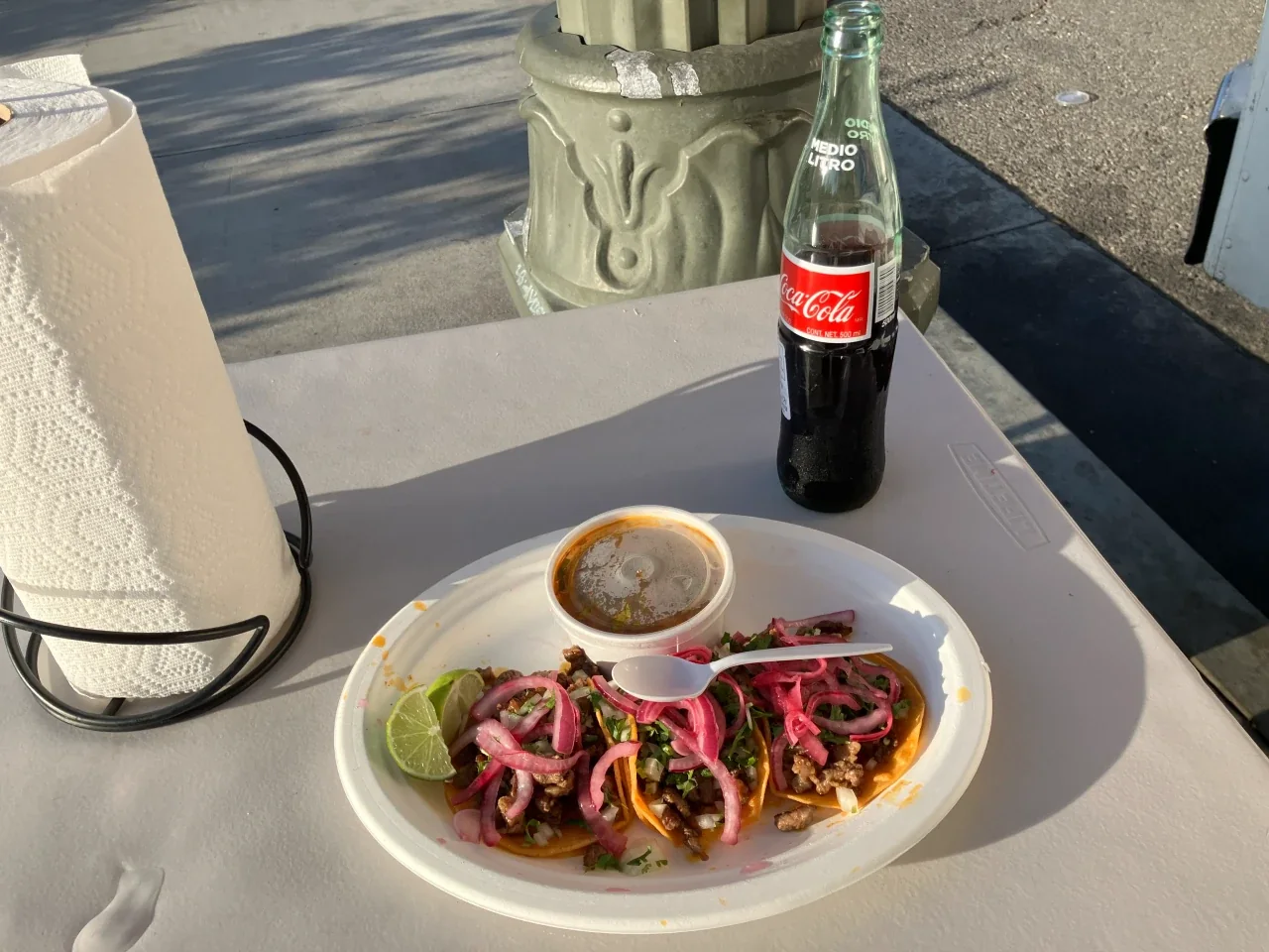 A plate of three Nikos Red Tacos with consommé and a Coca-Cola on an outdoor table